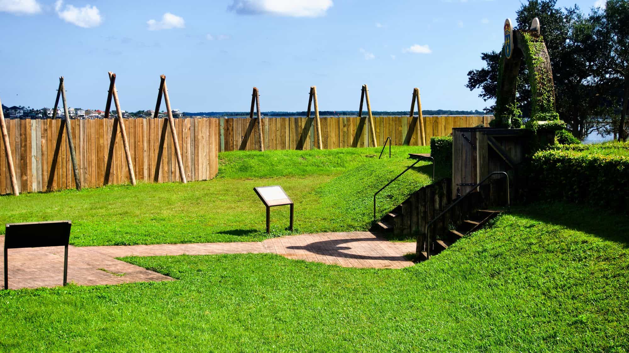 A reconstructed wooden fort wall and interpretive signs mark Fort Caroline National Memorial, with manicured green grass and views of the St. Johns River.