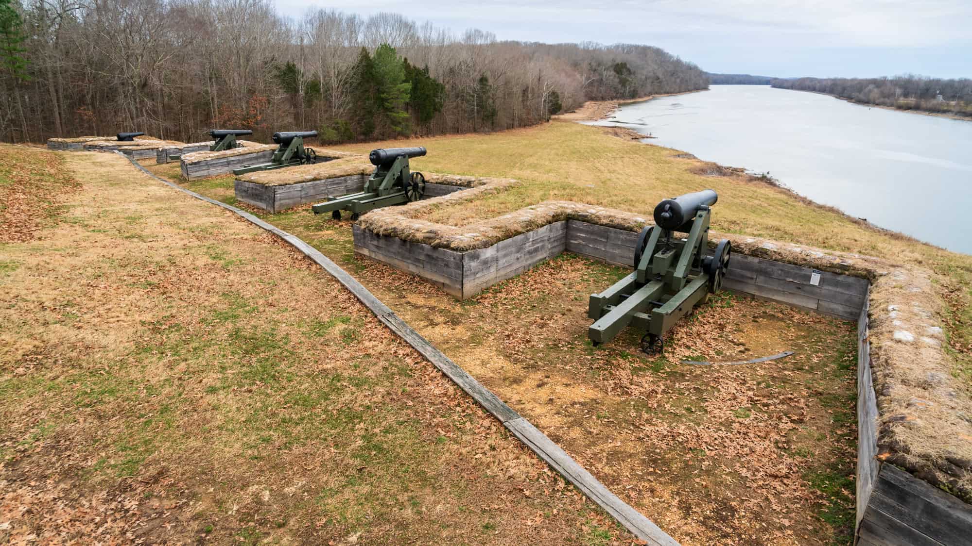 A row of historic Civil War-era cannons overlooks a river from raised wooden fortifications at Fort Donelson National Battlefield, surrounded by leaf-covered terrain.