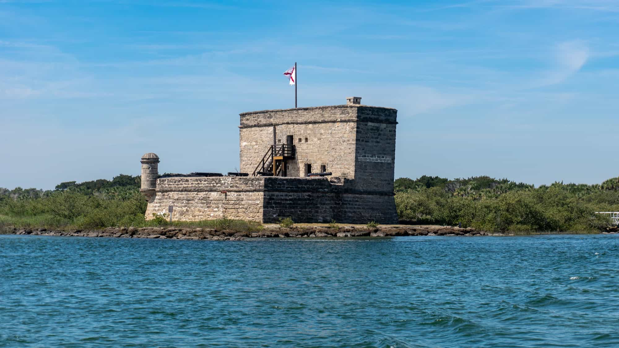 The small, historic Fort Matanzas sits on a marshy shoreline under a blue sky, with a red-crossed colonial Spanish flag flying above its stone walls.