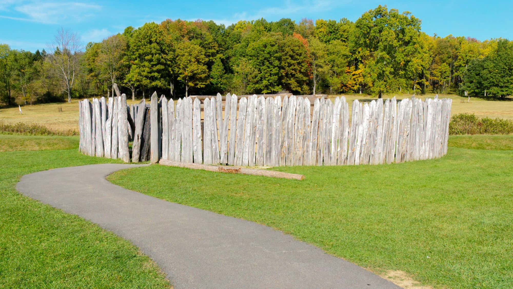 A circular wooden stockade fort built with tall, sharpened logs stands on an open grassy field, bordered by a paved path, with autumn-colored trees in the background.
