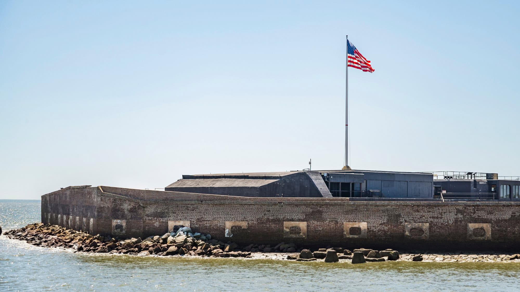 A low-profile, historic brick fort sits along the shoreline, partially surrounded by rocks, with an American flag waving from a tall flagpole under a clear blue sky.