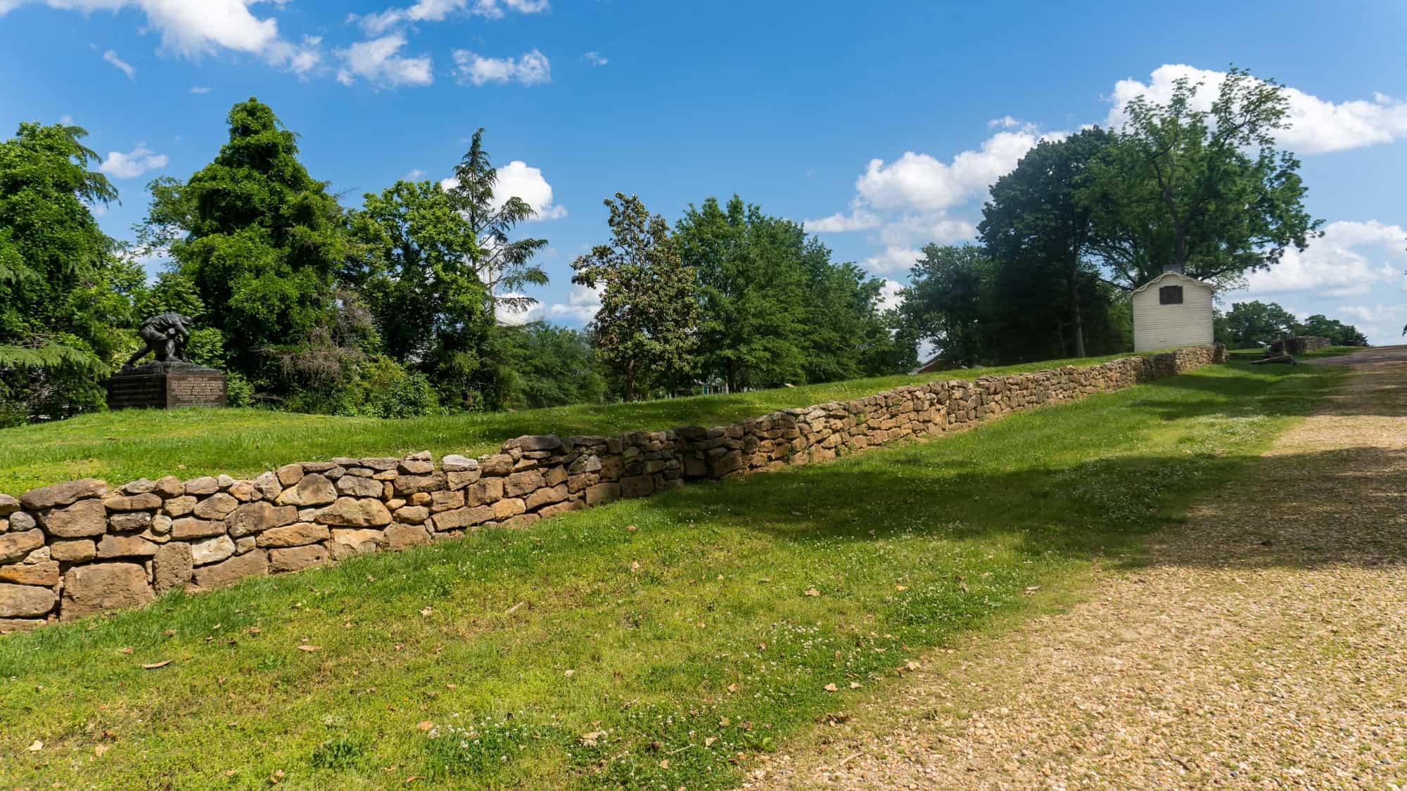 A low stone wall runs through Fredericksburg National Military Park, leading past a statue and toward a small white structure under a blue sky.
