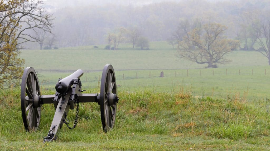 A solitary Civil War cannon on wooden wheels is positioned on a grassy hilltop, facing a misty green valley filled with scattered trees and surrounded by a wooden fence.