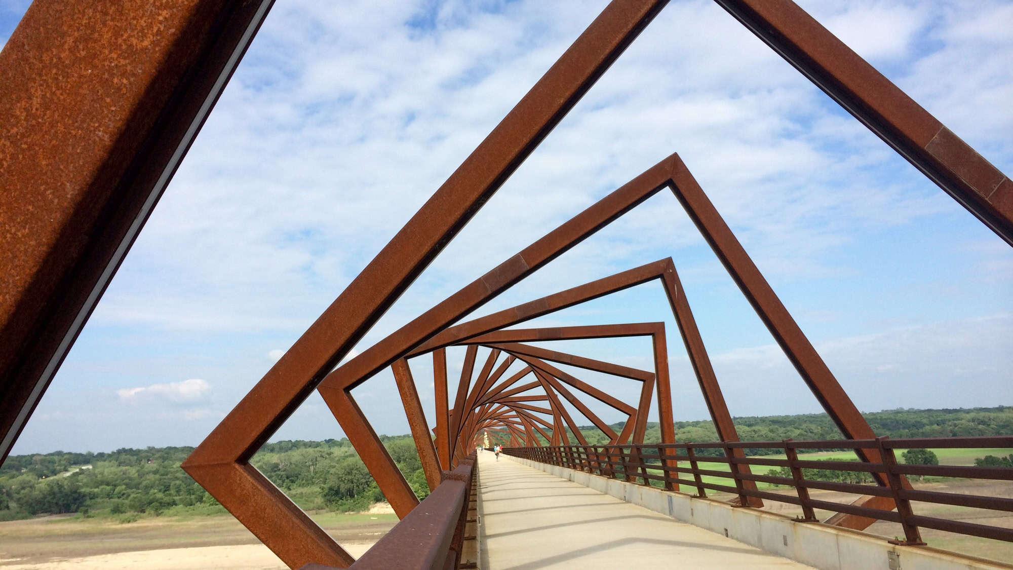 Striking geometric design of the High Trestle Trail Bridge, with rust-colored steel frames twisting over a wide path that spans a river valley, surrounded by blue skies and distant treetops.