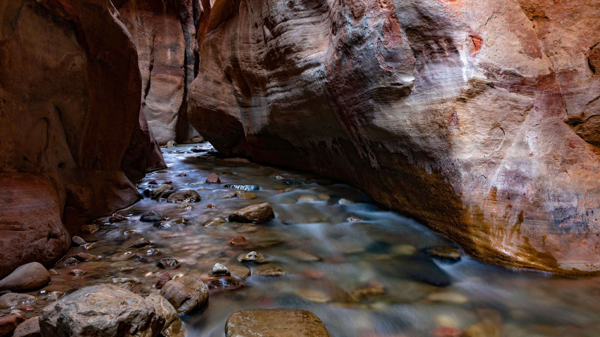 A narrow canyon with layered red rock walls and a gently flowing creek lined with smooth stones. The lighting highlights the natural textures and earthy tones of the slot canyon.