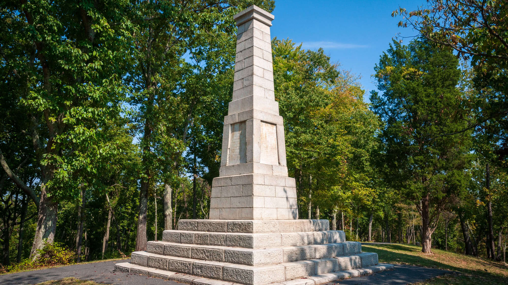 A tall stone obelisk monument stands on a platform in Kings Mountain National Military Park, framed by dense green trees and a clear sky.