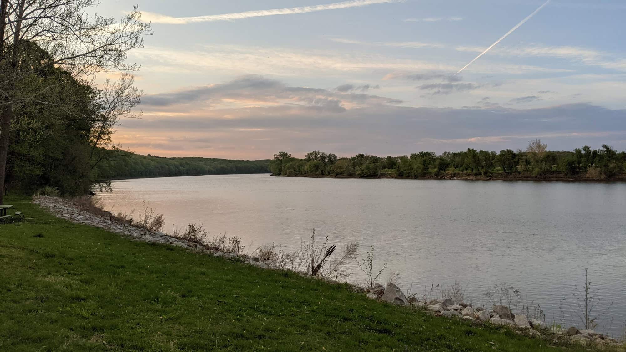 Peaceful scene of a river or lake at sunset with a grassy bank and rocky shoreline in the foreground, trees lining both sides of the water, and pastel-colored clouds streaked across the sky.
