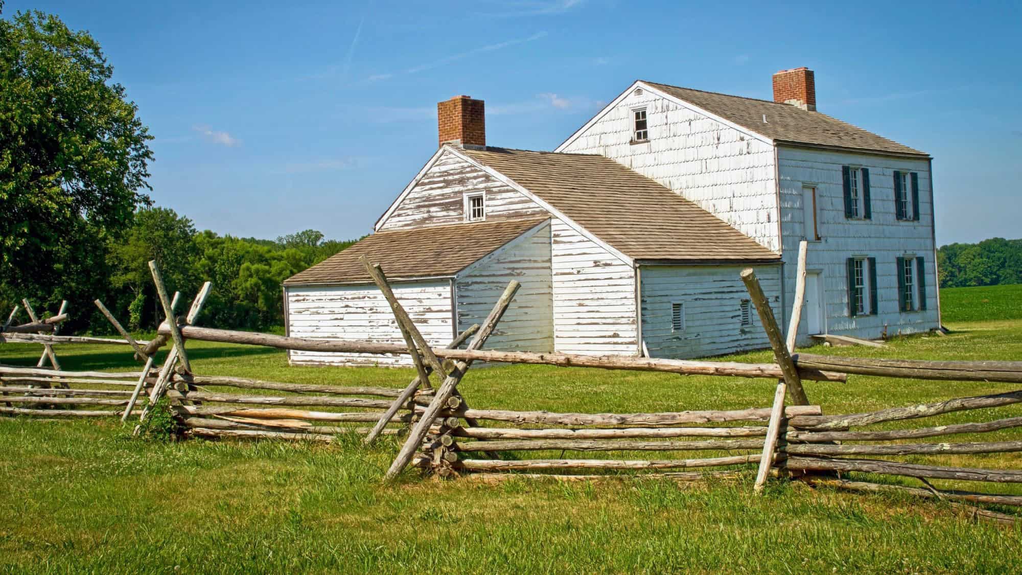 A weathered white farmhouse with two chimneys and a wood-shingled roof sits behind a split-rail fence at Monmouth Battlefield State Park, surrounded by open grassy fields.