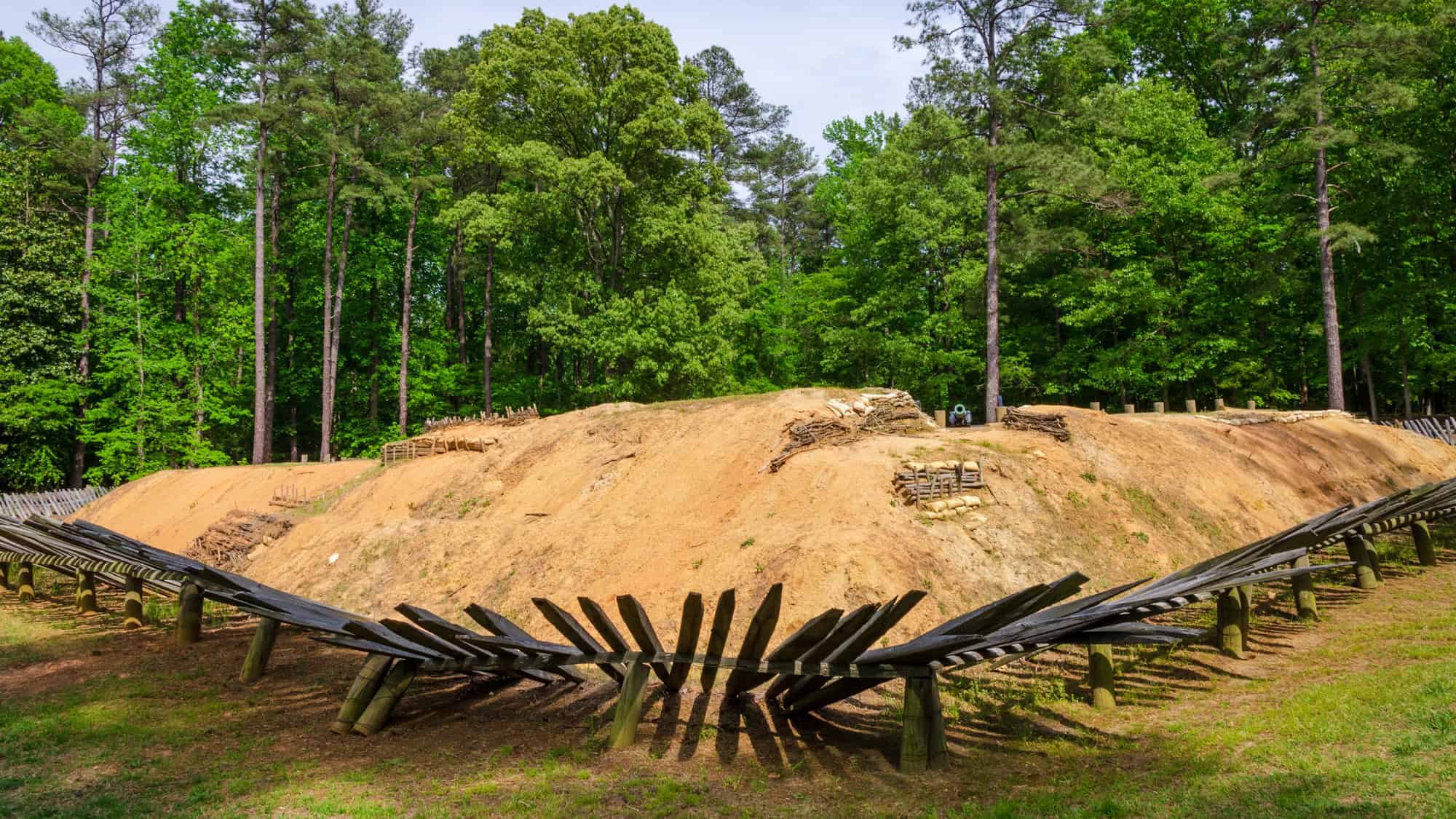 A restored Civil War-era earthwork fortification surrounded by dense forest, with sharply angled wooden chevaux-de-frise defenses in the foreground and sandbag-lined trenches visible along the embankment.