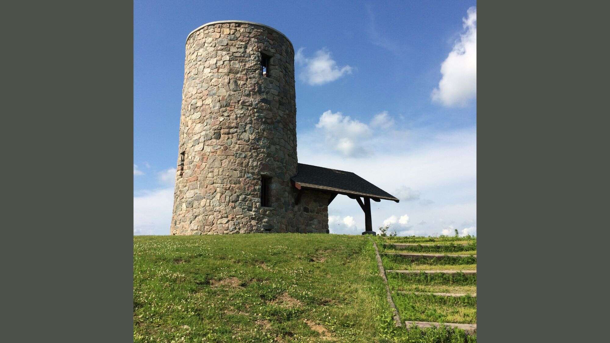 Round stone tower with small windows sits atop a grassy hill under a bright blue sky with puffy white clouds, beside a short stairway leading up to it.