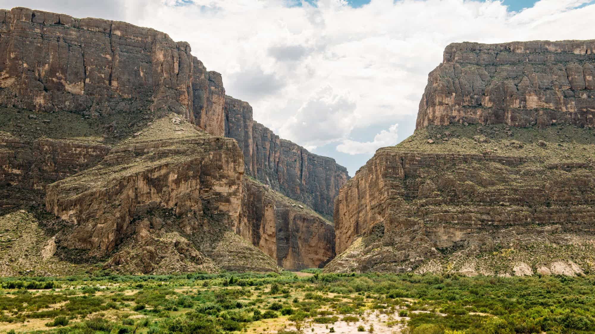 A dramatic view of Santa Elena Canyon, where the Rio Grande snakes between two towering rock faces. The flat, green valley floor contrasts with the sheer cliffs above.