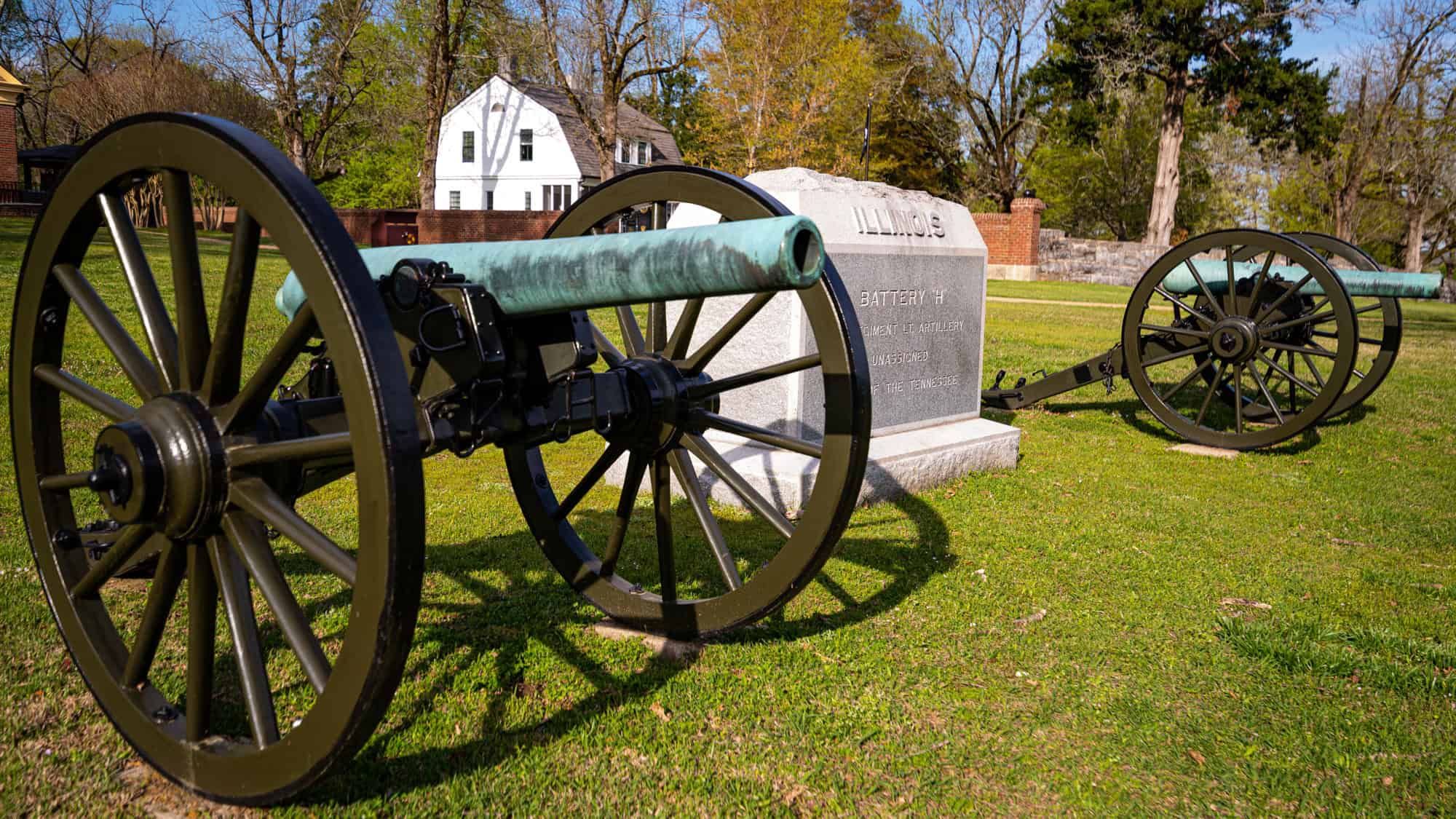 Two historic cannons flank a granite memorial inscribed with "ILLINOIS" at Shiloh National Military Park, surrounded by lush green grass and historic buildings in the background.