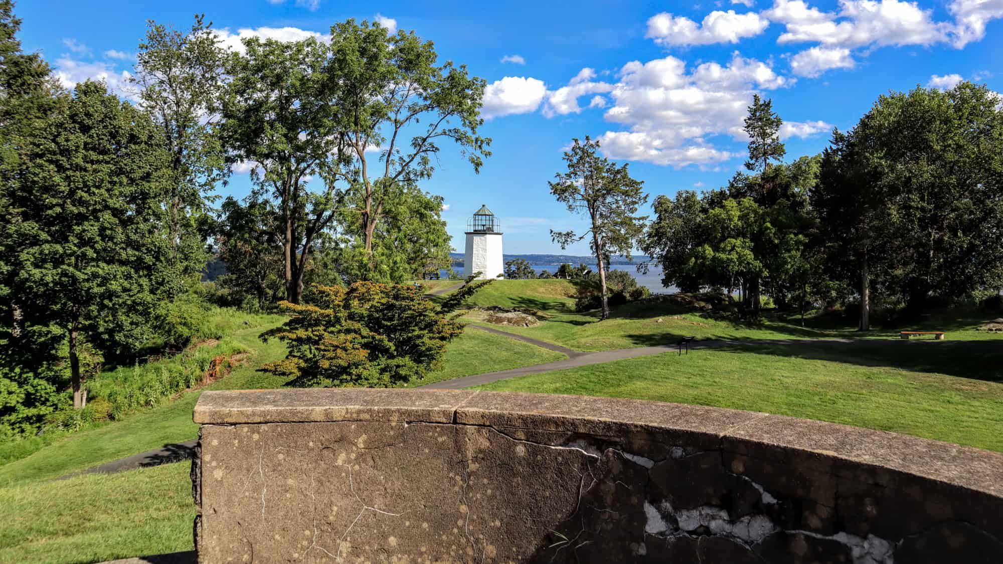A small white lighthouse with a glass-topped lantern rises above the trees in Stony Point Battlefield, overlooking the Hudson River on a bright, partly cloudy day.