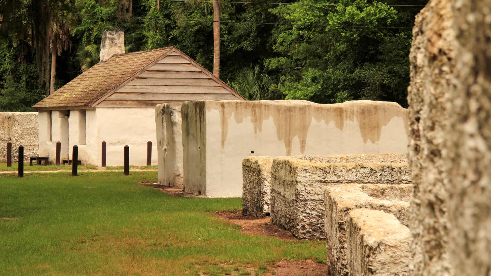 Historic coquina stone walls and a reconstructed tabby cabin sit in a shaded clearing, surrounded by grass and forest in the Timucuan Preserve.