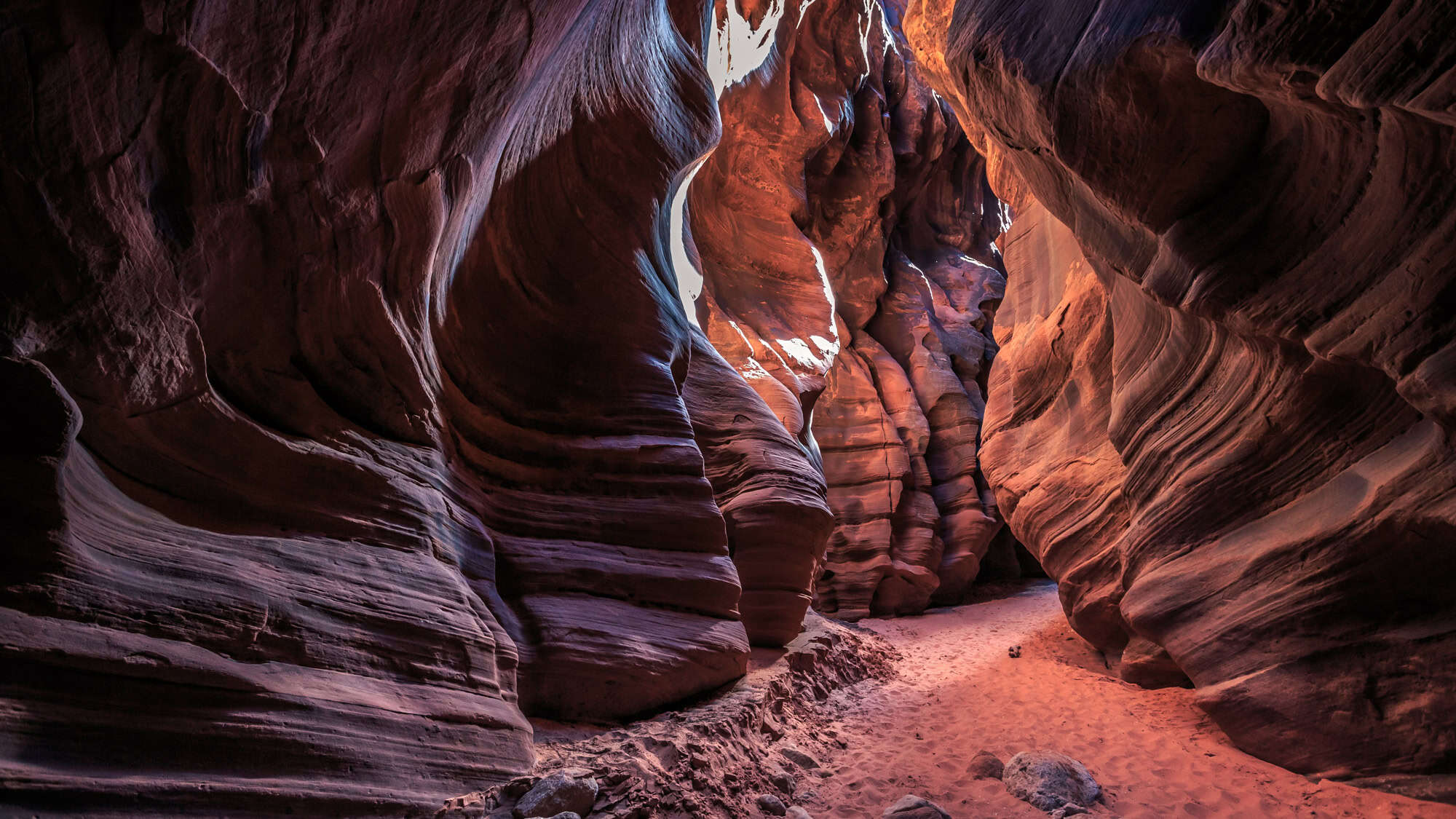 A winding slot canyon with dramatically curved, striated red rock walls and a sandy path, illuminated by soft natural light from above.