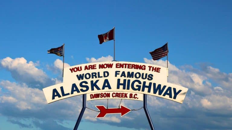 A bright white and red sign welcomes travelers to the "World Famous Alaska Highway" in Dawson Creek, British Columbia, with the U.S., Canadian, and British Columbia flags waving above against a clear blue sky.