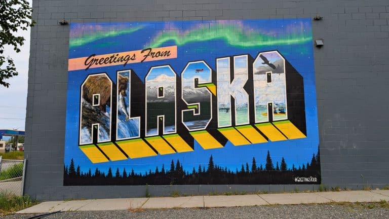 A vibrant mural on a gray wall reads “Greetings from Alaska,” with each letter of “ALASKA” filled with scenic imagery—grizzly bears, mountains, glaciers, northern lights, and an eagle soaring—celebrating the state’s wild beauty. The mural pops with bold yellow shadows and silhouettes of pine trees beneath a vivid blue and green aurora.