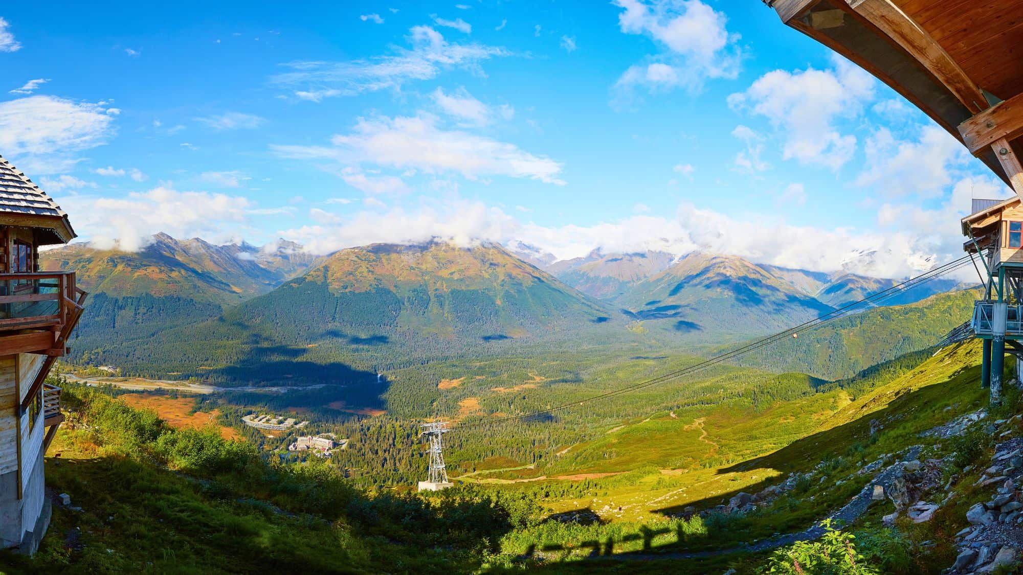 A sweeping panoramic view from the Alyeska Resort shows green forested slopes, alpine meadows, and rugged mountain peaks under a clear blue sky, with low clouds clinging to the ridges. Aerial tram cables stretch across the scene, linking the mountainside lodge buildings perched at the edges.