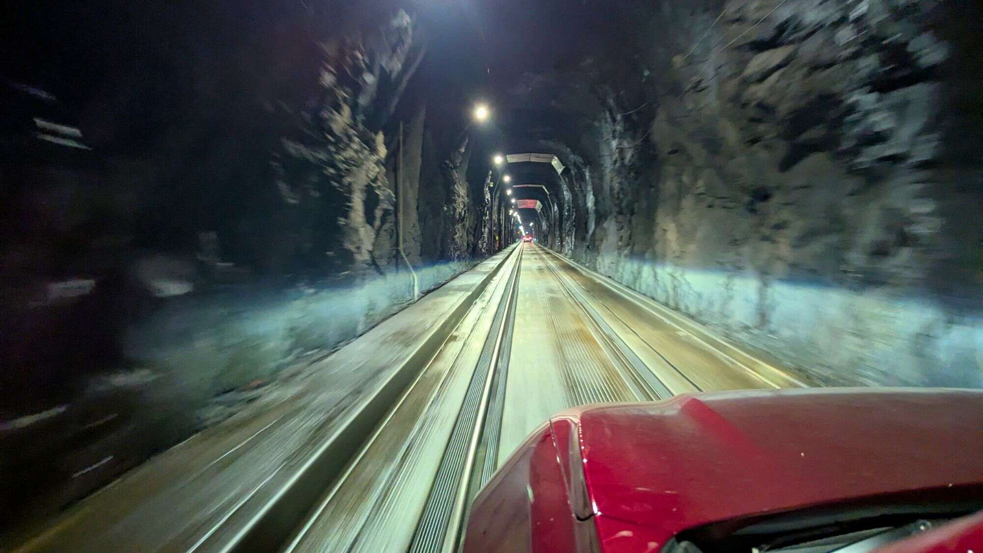 View from a red car driving through the Anton Anderson Memorial Tunnel, a long, narrow one-lane tunnel with rugged rock walls, train tracks down the center, and overhead lights casting a dramatic glow. The perspective emphasizes the tunnel's depth and engineered path through solid mountain rock.