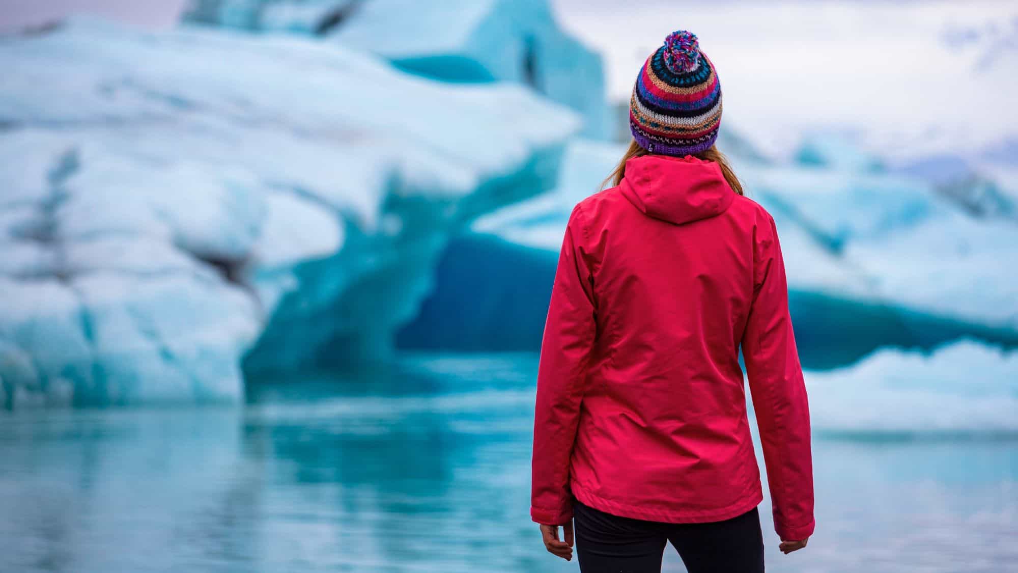 A woman in a red jacket and colorful knit hat stands at the edge of a calm glacial lake, gazing at massive blue ice formations in the distance.