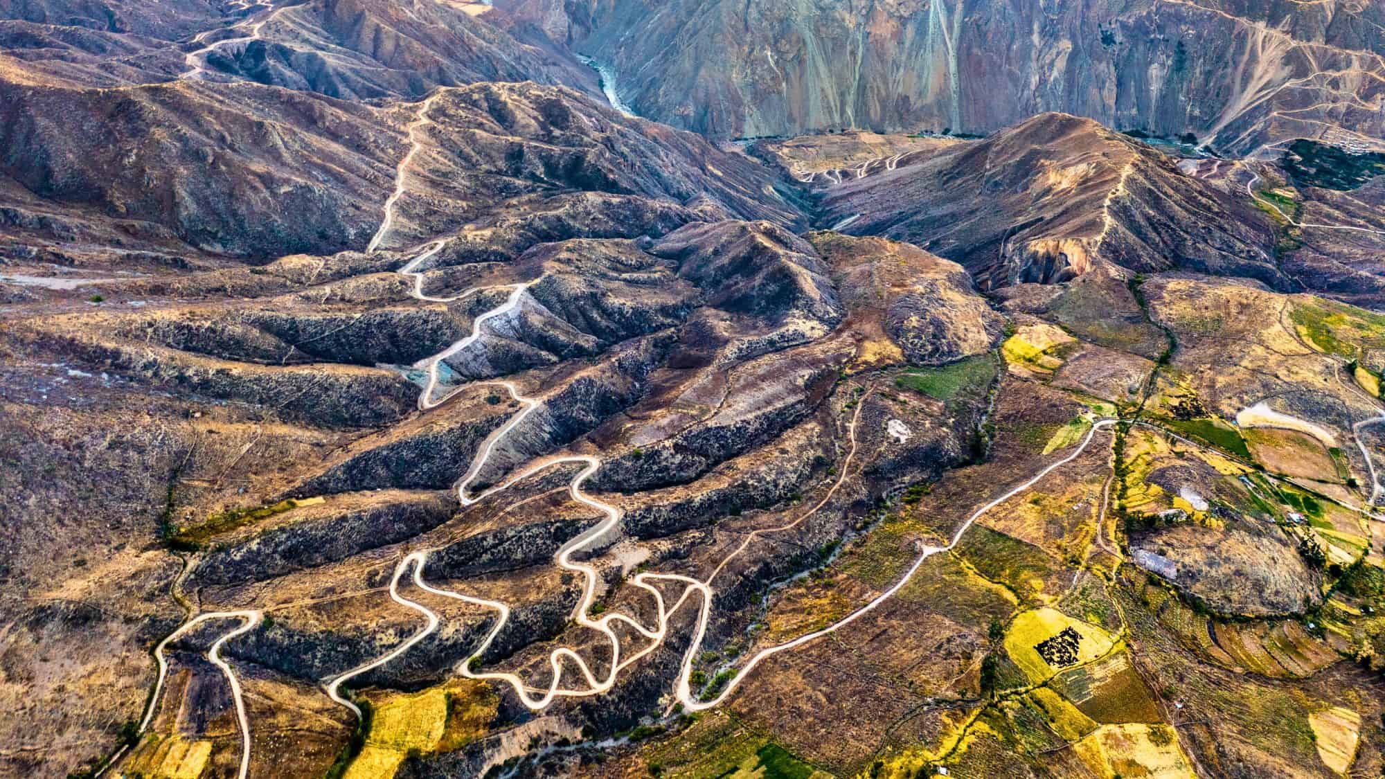 A high-altitude aerial view of a winding dirt road snaking through rugged brown Andean terrain dotted with fields and deep valleys.