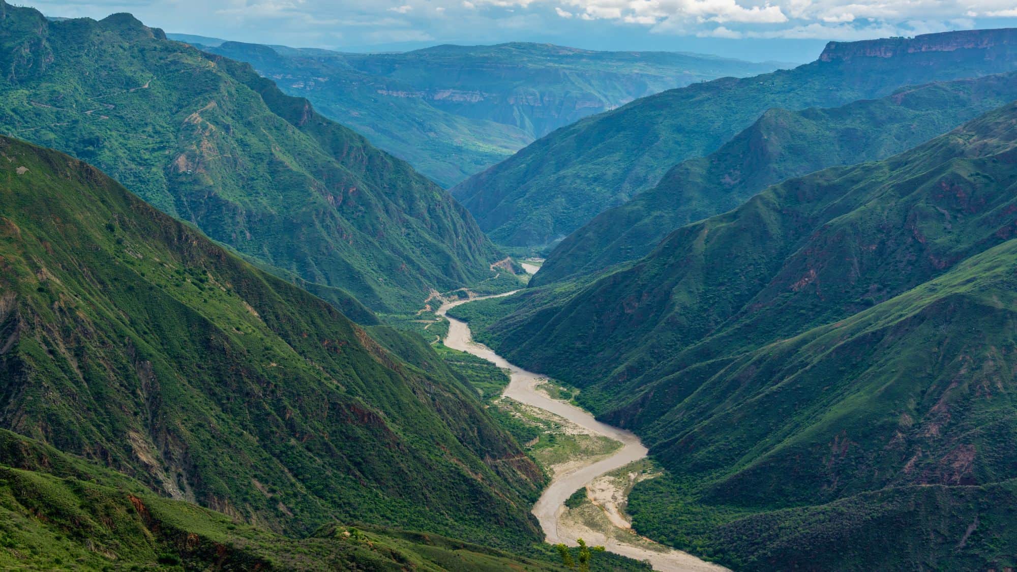 A lush, steep canyon blanketed in greenery with a winding river flowing through the valley floor under a partly cloudy sky.