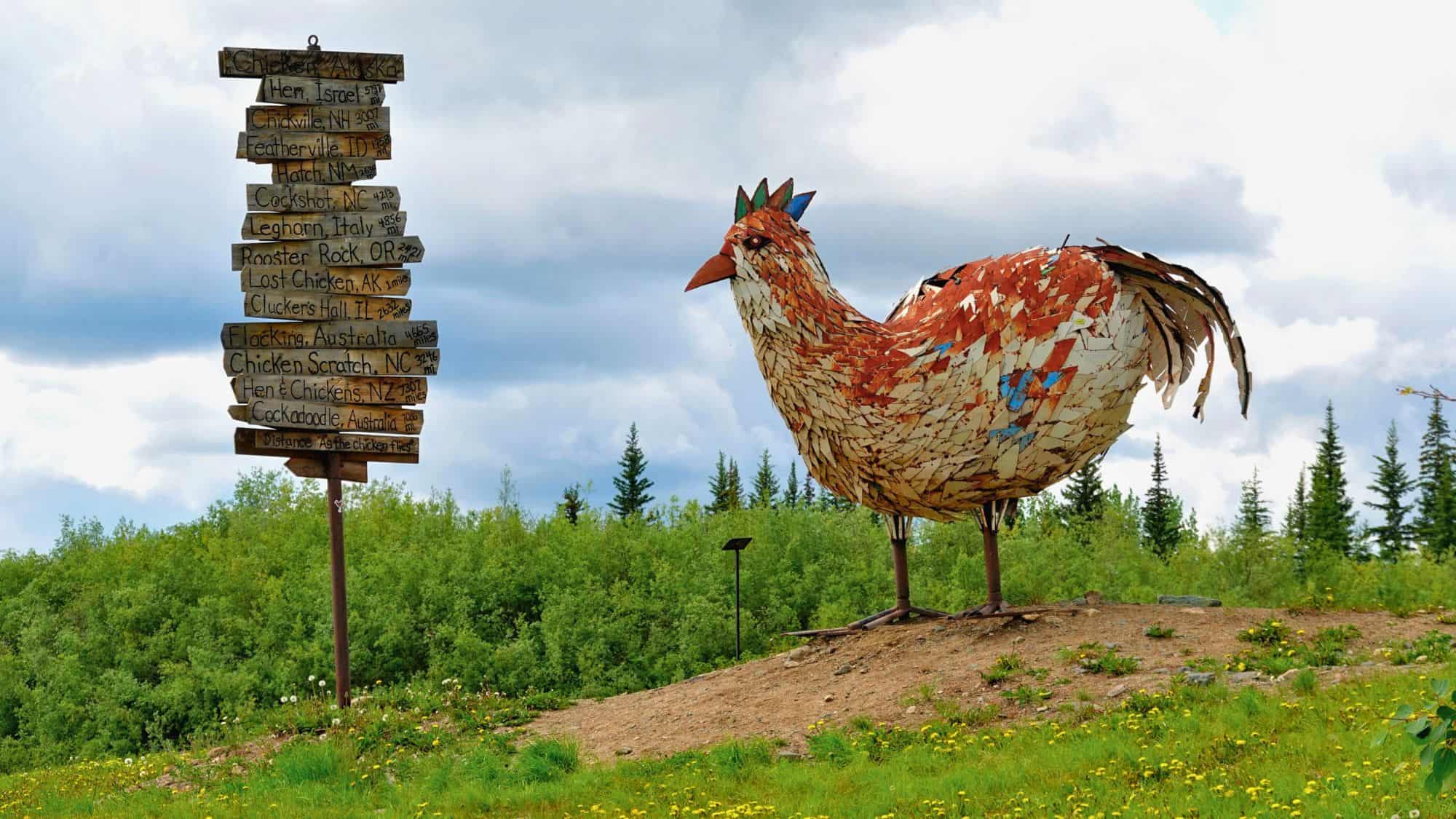A quirky roadside attraction in Chicken, Alaska, features a massive rust-colored chicken sculpture made from metal panels, next to a whimsical signpost pointing to other poultry-themed destinations around the world.
