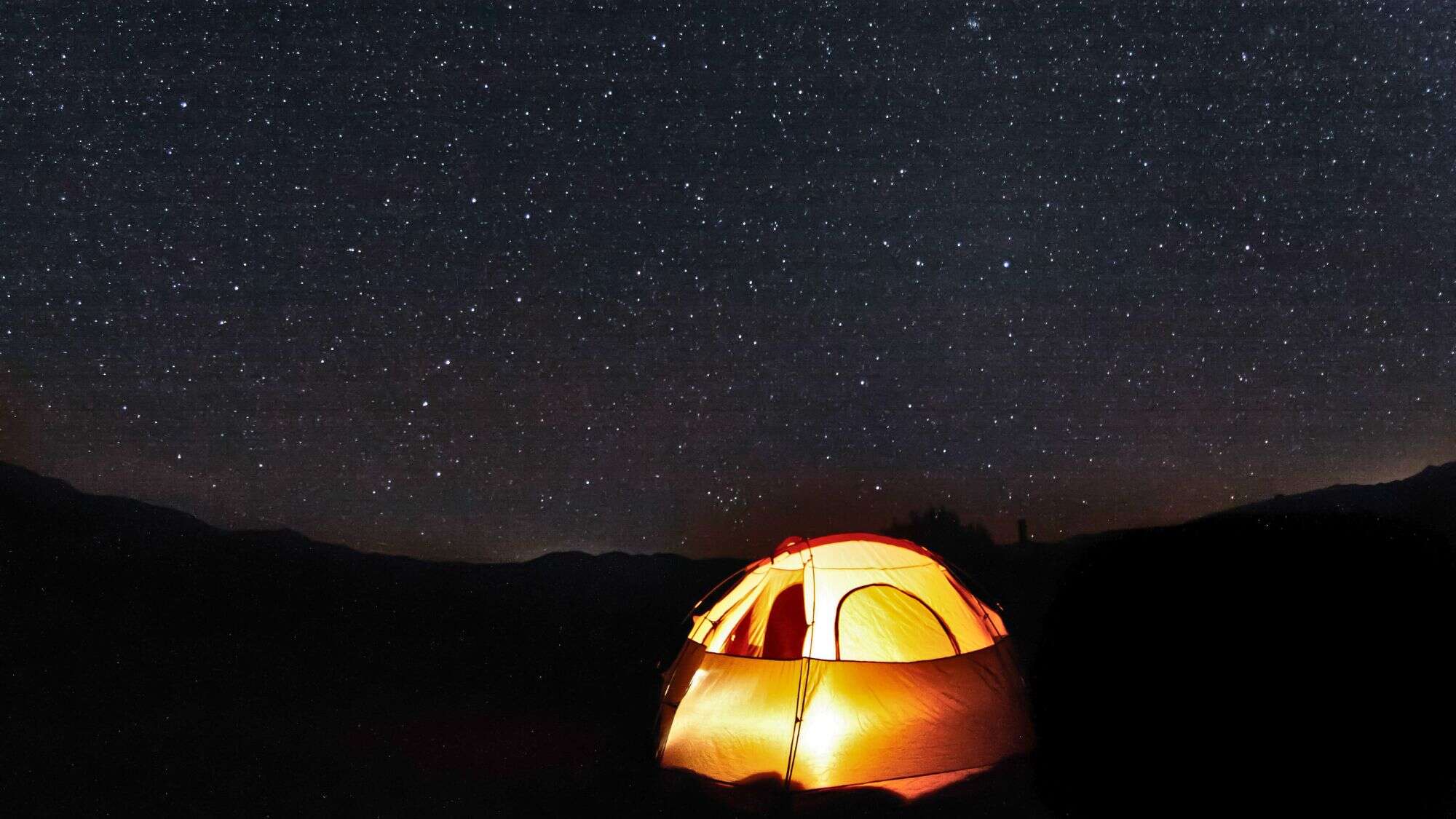 A warmly lit tent glows under a sky bursting with stars, set against the silhouette of distant mountains, capturing the peaceful solitude of a remote night under the stars.