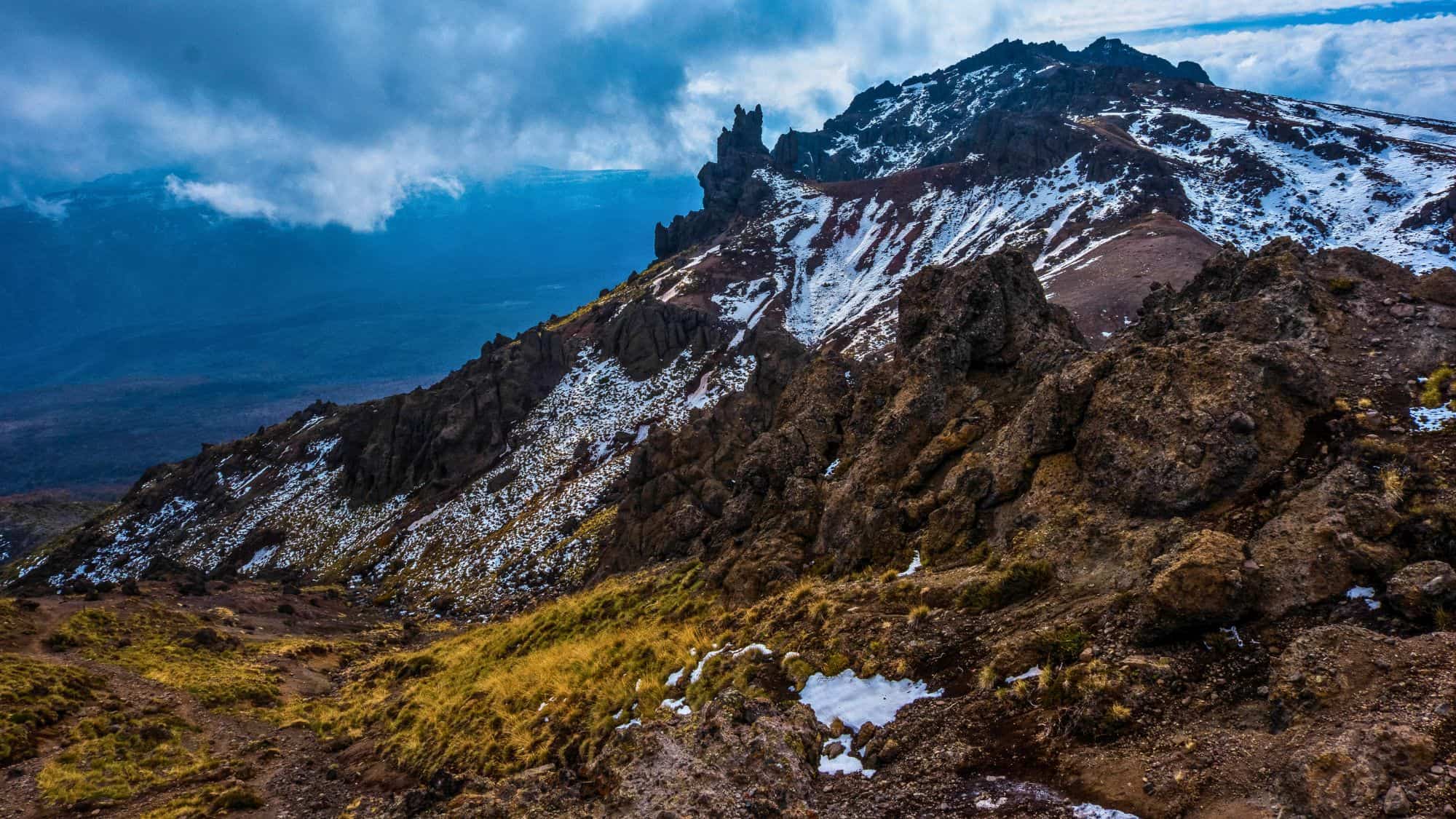Jagged, rocky terrain leads to dramatic volcanic peaks partially covered in snow, with clouds drifting over a moody mountain landscape.