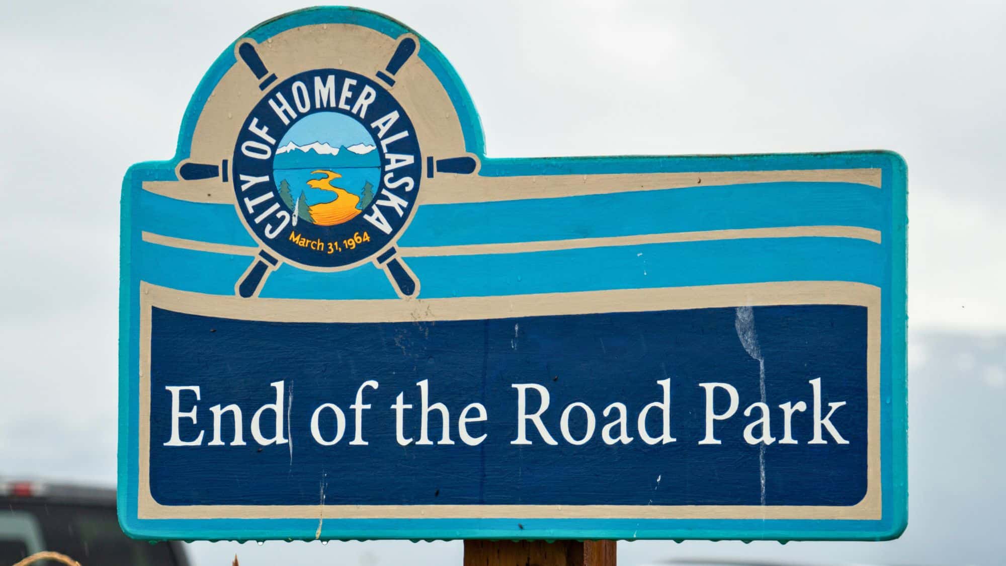 A brightly painted wooden sign reads “End of the Road Park” in Homer, Alaska, with a nautical wheel and scenic logo, marking a coastal milestone.