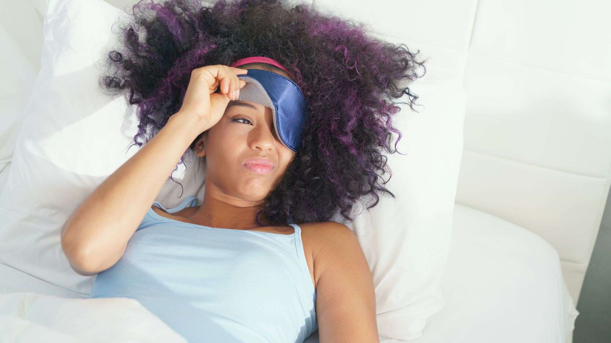 A woman lies in bed lifting a sleep mask with a groggy, unimpressed expression, her curly hair spilling over white bedding and a blue tank top.