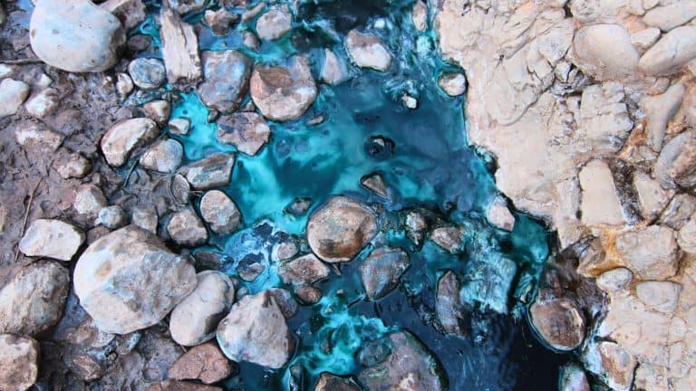 A vivid, close-up image of turquoise and deep blue geothermal water flowing between smooth, rounded rocks, highlighting the mineral-rich textures and colors.