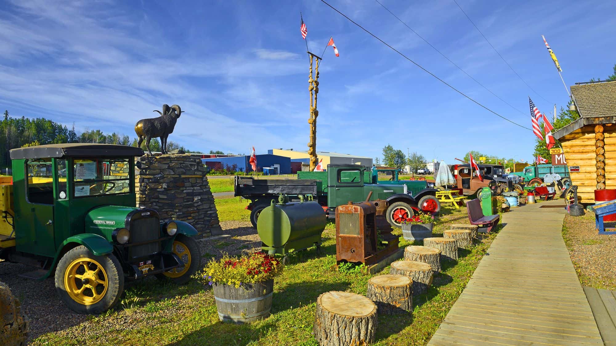 A vibrant outdoor display of antique cars, logging and mining equipment, and vintage trucks at the Fort Nelson Heritage Museum, with flags flying and a bronze ram statue atop a rock pedestal.