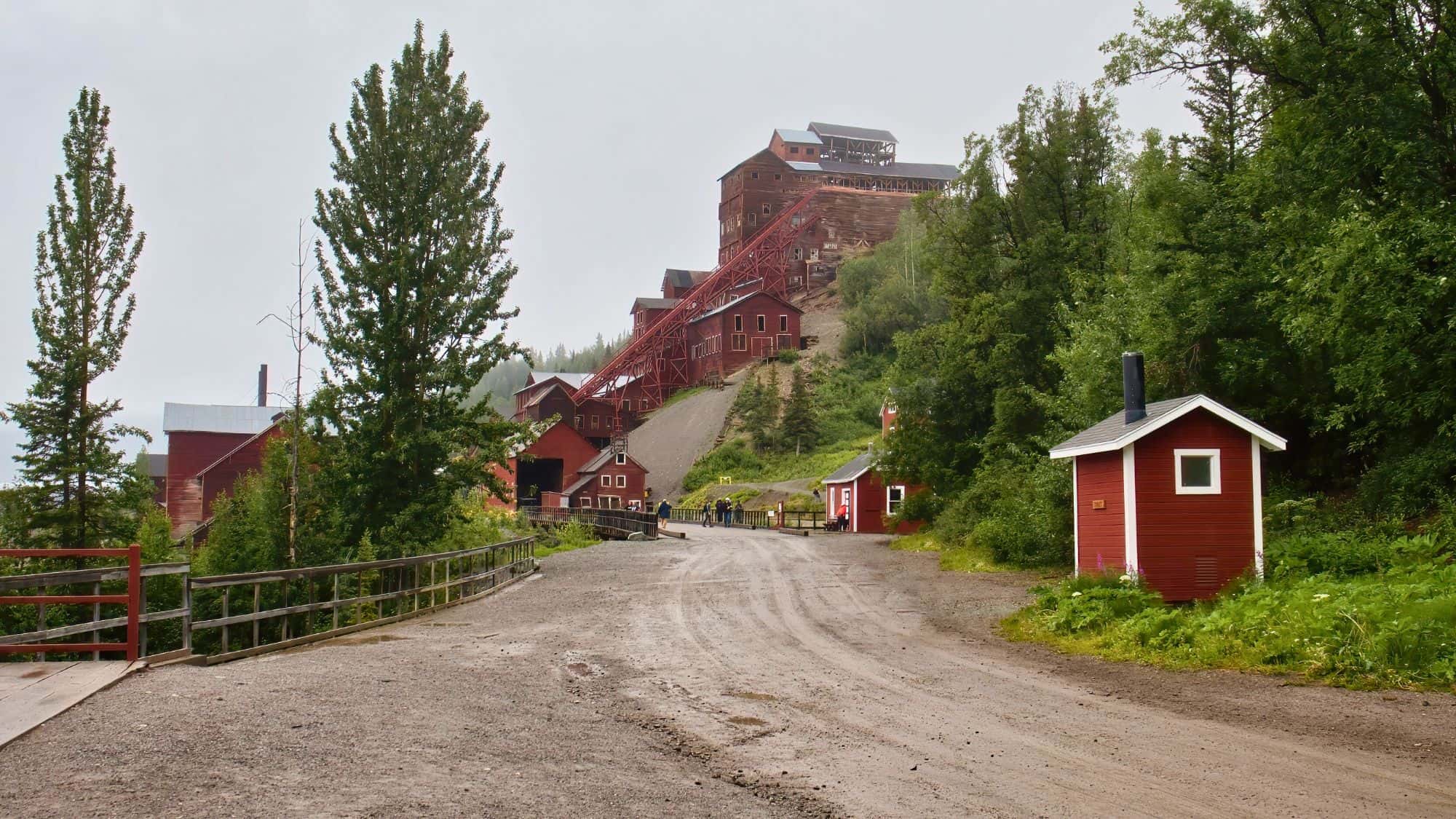 A historic red mining complex climbs a green hillside under cloudy skies in Kennecott, Alaska, with weathered buildings and a gravel road leading through the preserved ghost town.