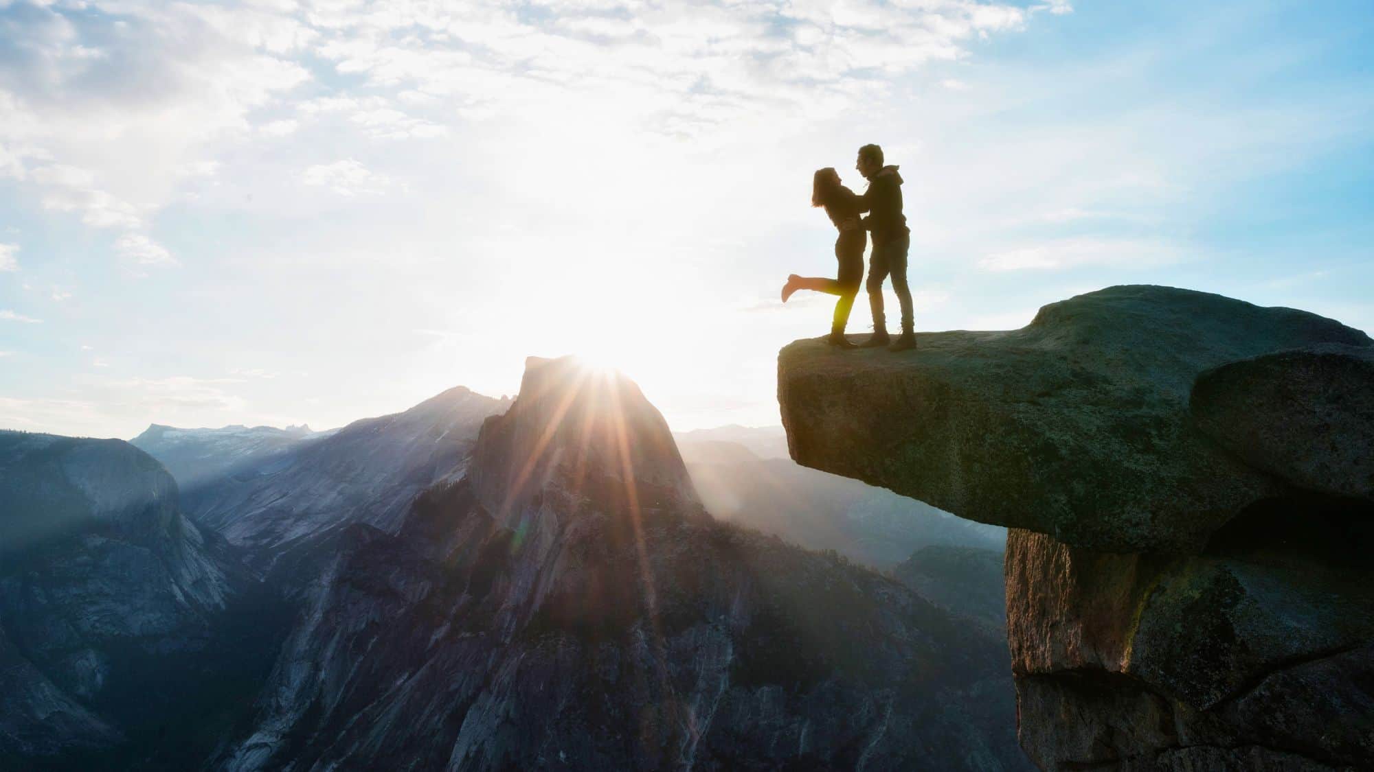 A silhouetted couple embraces on the edge of a dramatic rock outcropping with one person lifted off the ground, backlit by the rising sun over a mountainous landscape.