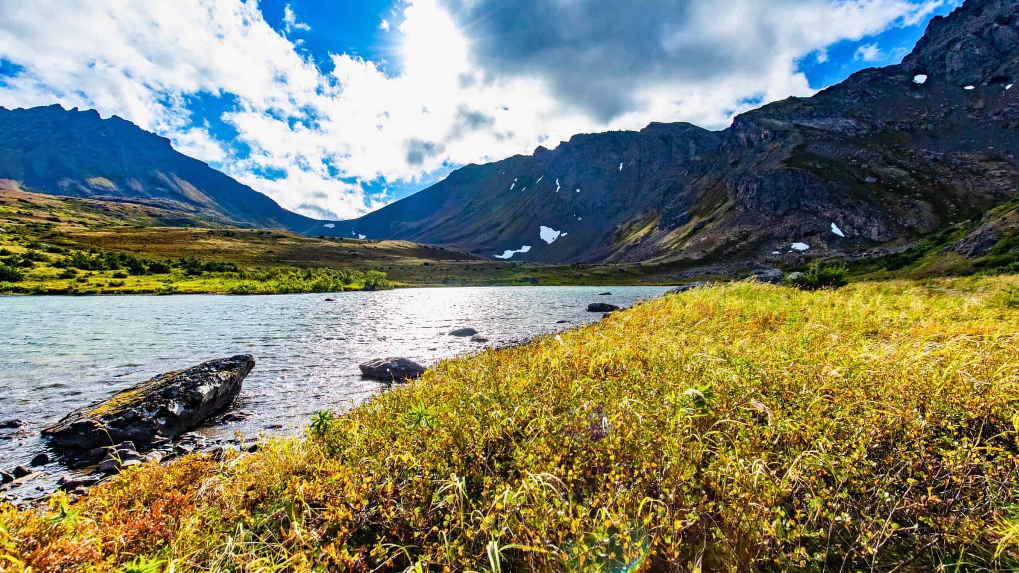 A sparkling alpine lake nestles at the base of steep, rugged mountains with patchy snow, while golden grasses sway in the foreground under a partly cloudy sky with dramatic lighting.