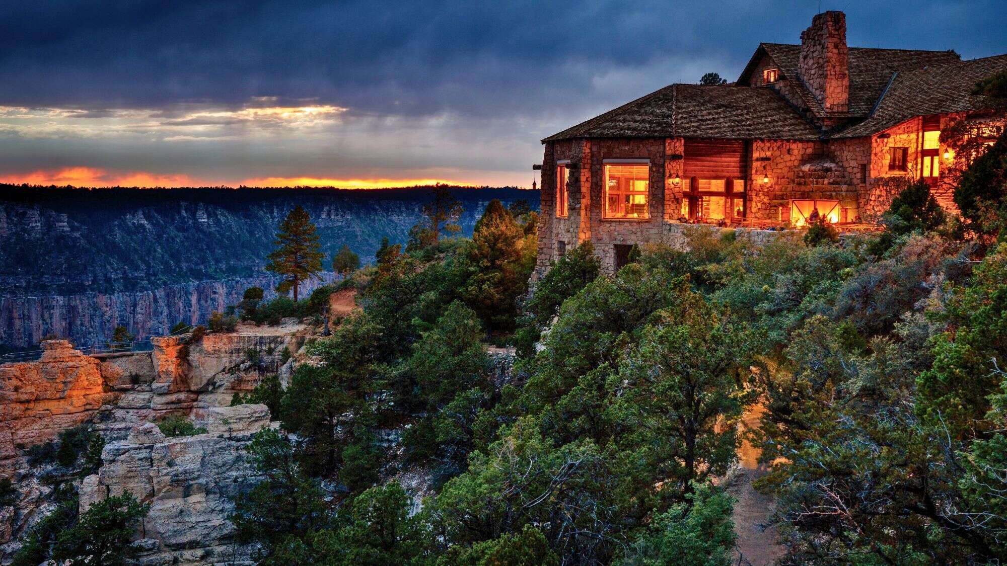 A warm, rustic stone lodge glows at the edge of the Grand Canyon’s North Rim, with dramatic cliffs and trees framing a fiery sunset sky.