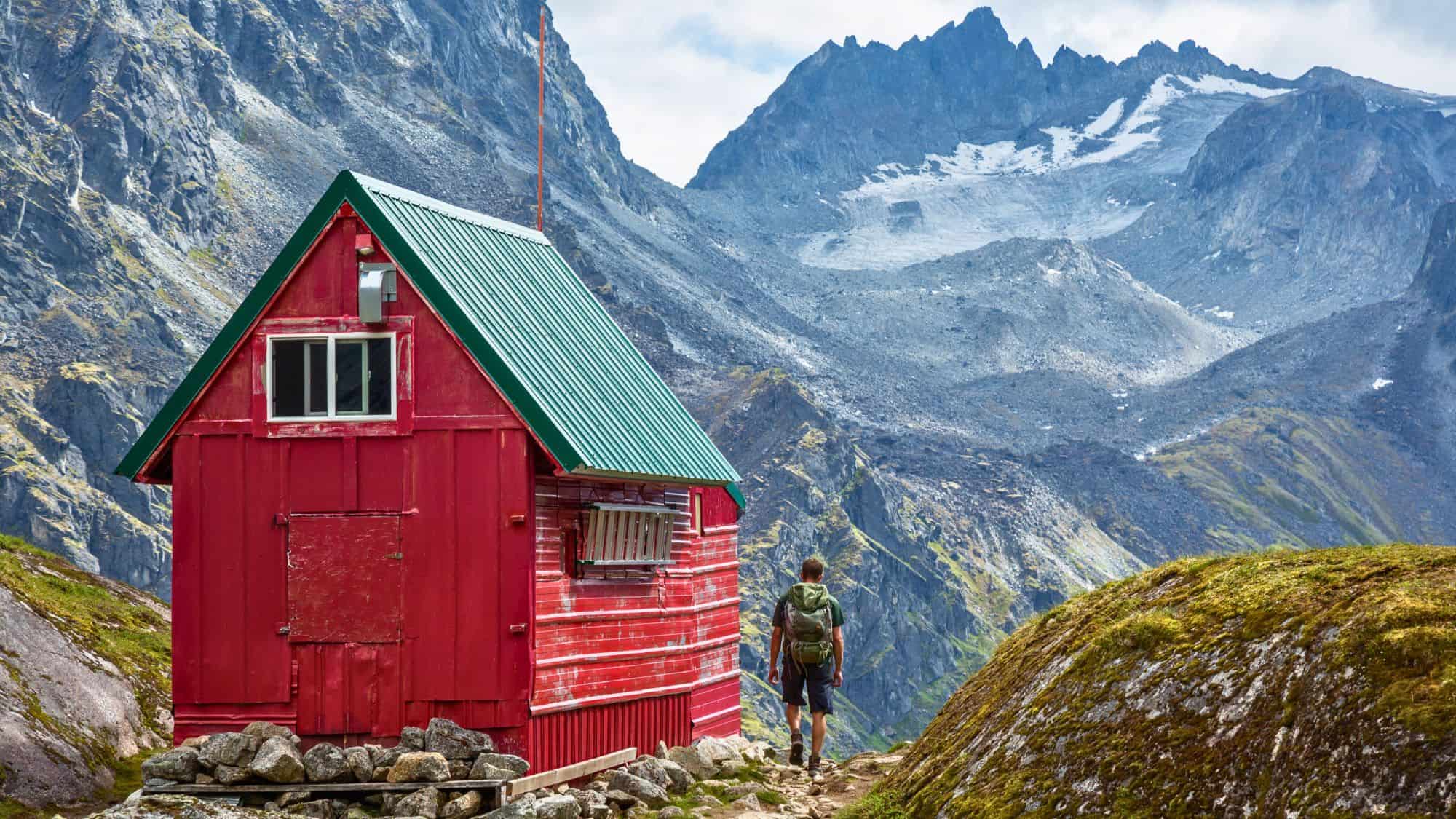 A hiker approaches a vibrant red backcountry cabin with a green roof, set against dramatic alpine peaks and glacier-carved ridges in Hatcher Pass, Alaska.