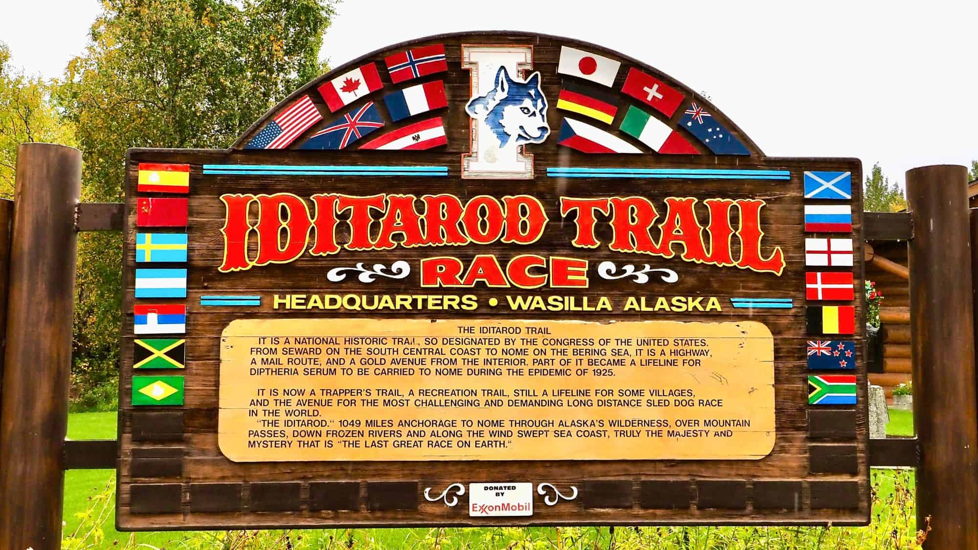 A large wooden sign at the Iditarod Trail Headquarters in Wasilla, Alaska, displays the race name flanked by colorful international flags. Text below describes the historical and modern significance of the famed sled dog race.