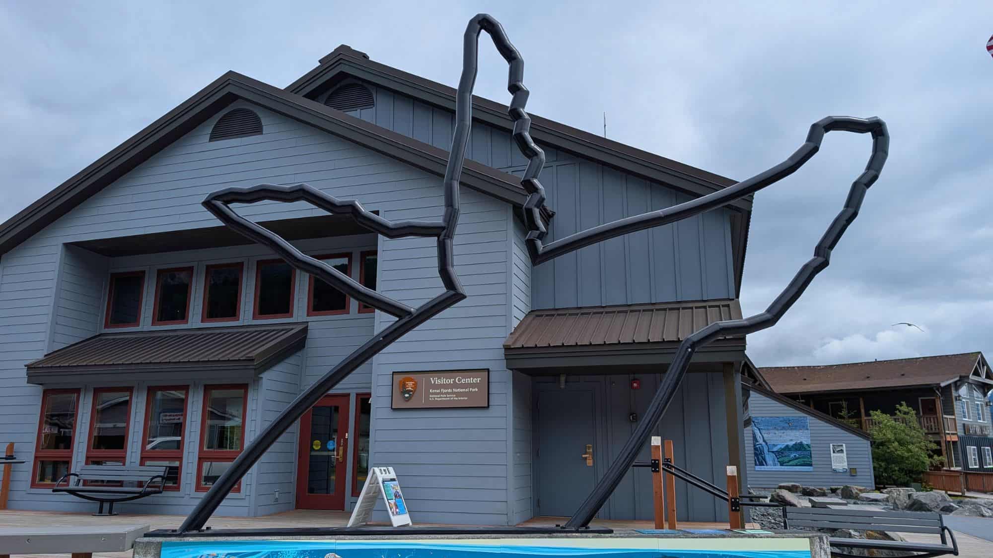 A large metal sculpture shaped like a map of Alaska stands in front of the Kenai Fjords National Park Visitor Center, a gray and red building with NPS signage and informational displays outside.