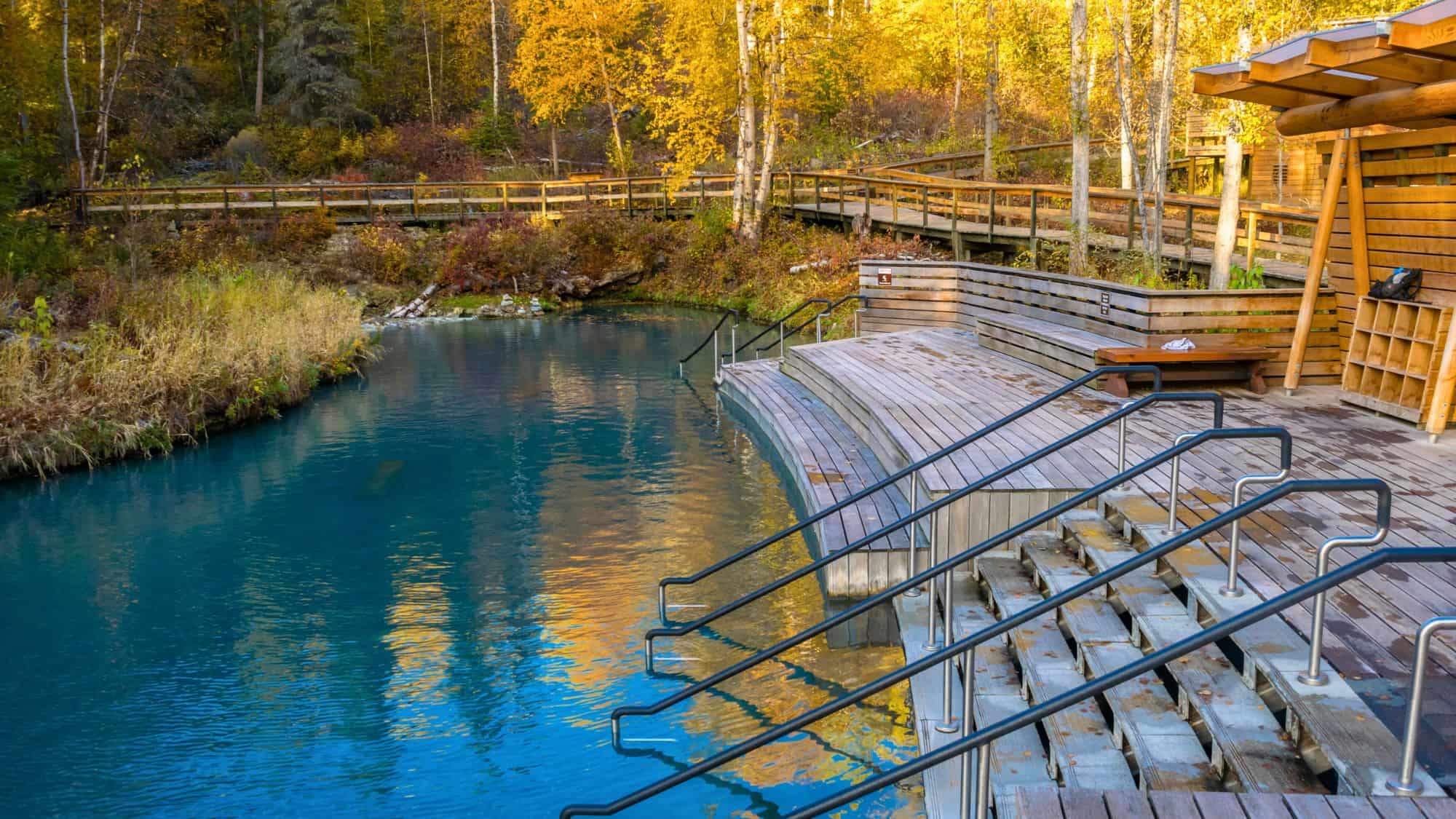 A serene hot springs pool with teal blue water and tiered wooden steps, set in a lush forest with golden autumn foliage and boardwalks winding through the trees at Liard River Hot Springs Provincial Park.