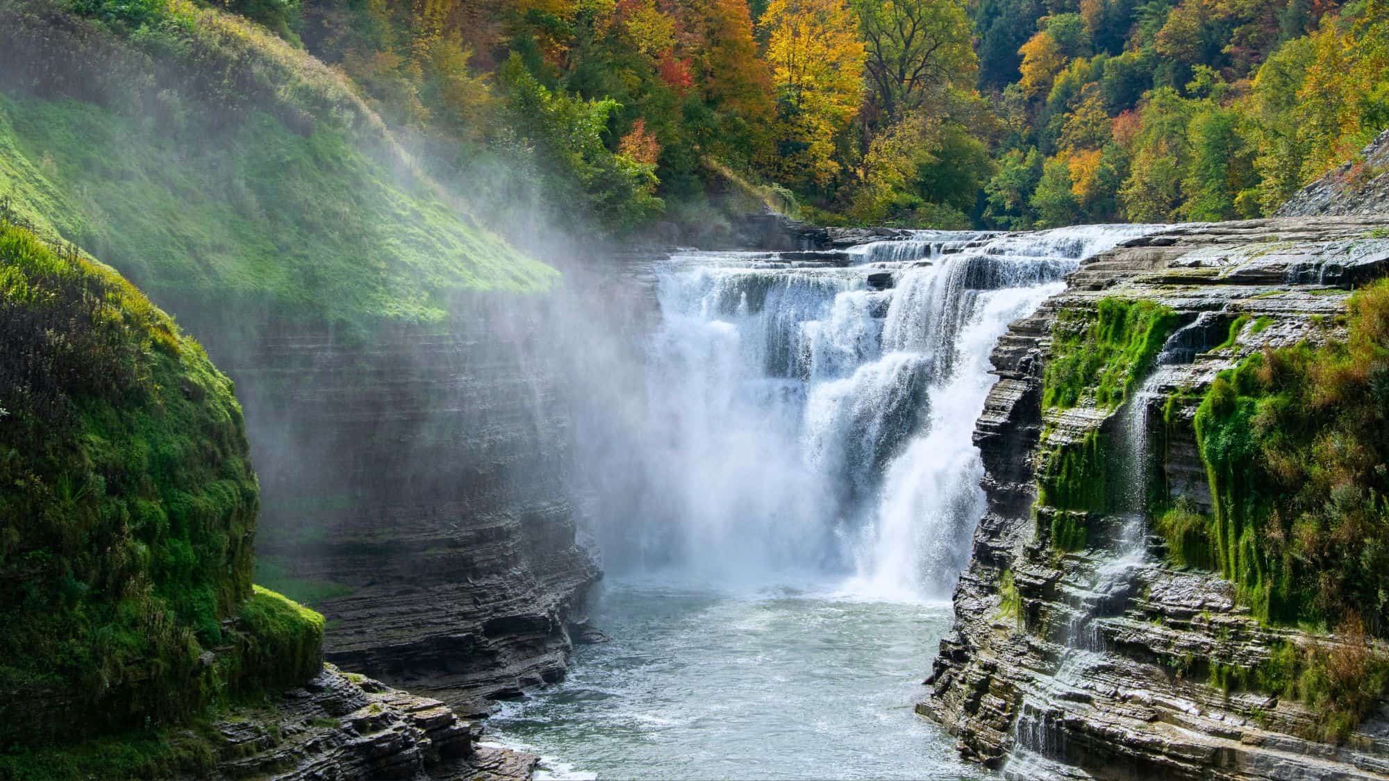 A wide, roaring waterfall spills over layered rock ledges into a gorge surrounded by green moss and colorful autumn foliage, with mist rising in the air.