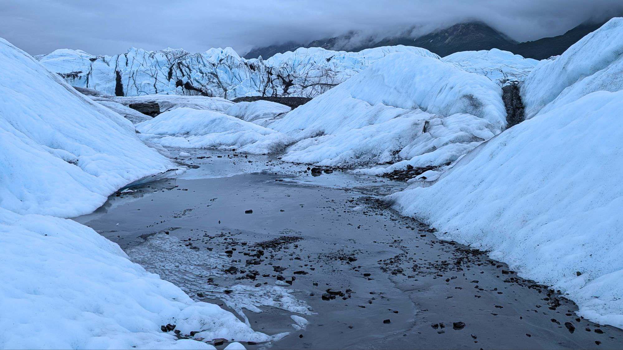 Icy blue glacier ridges rise on either side of a frozen, gravel-strewn meltwater channel, with low clouds obscuring the mountains in the background.