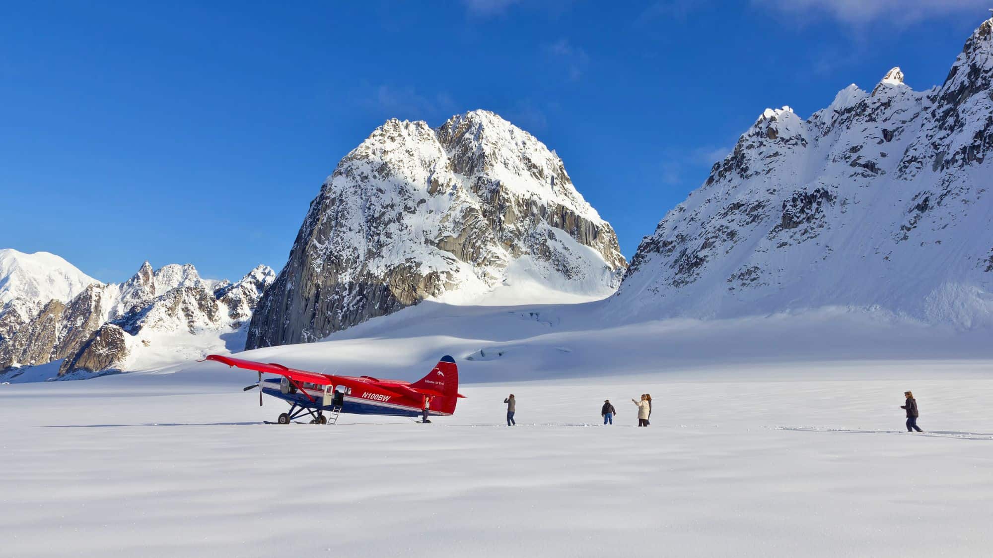 A red bush plane rests on a vast, snowy glacier surrounded by jagged alpine peaks, as bundled-up people walk across the frozen expanse under a clear blue sky.