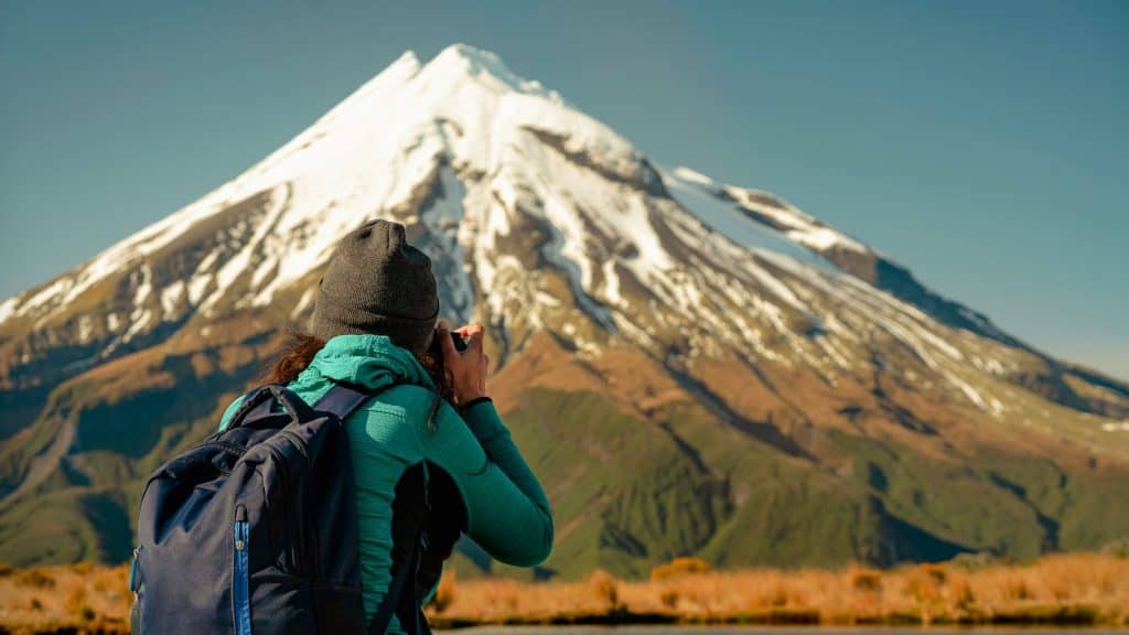 A woman in outdoor gear and a backpack photographs Mount Taranaki’s snow-draped slopes from a close, elevated viewpoint in the morning light.