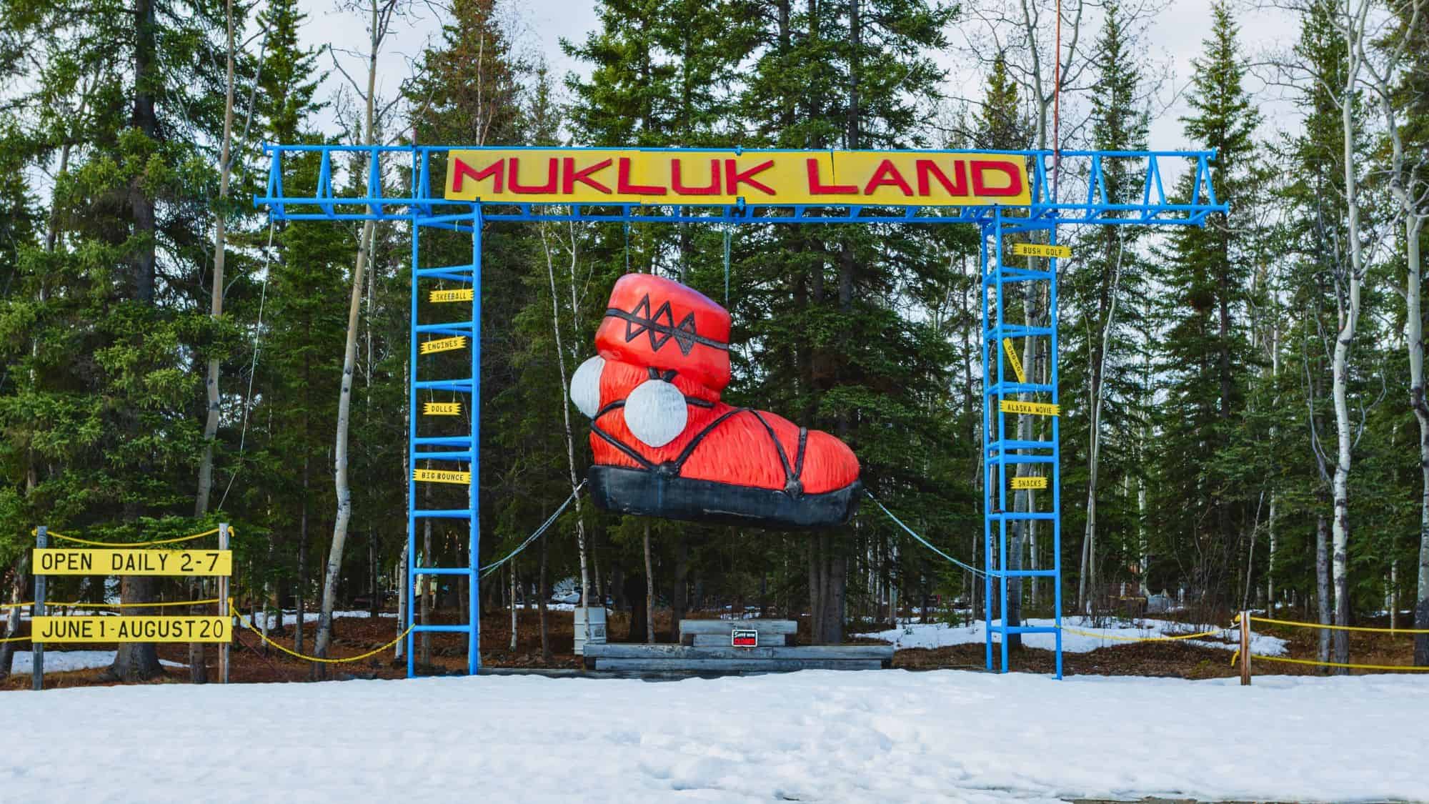 A whimsical roadside attraction sign for Mukluk Land features a giant red mukluk boot suspended from a bright blue metal frame, surrounded by snow and pine trees, with signs advertising skeeball, engines, dolls, snacks, and an “Alaska movie.”