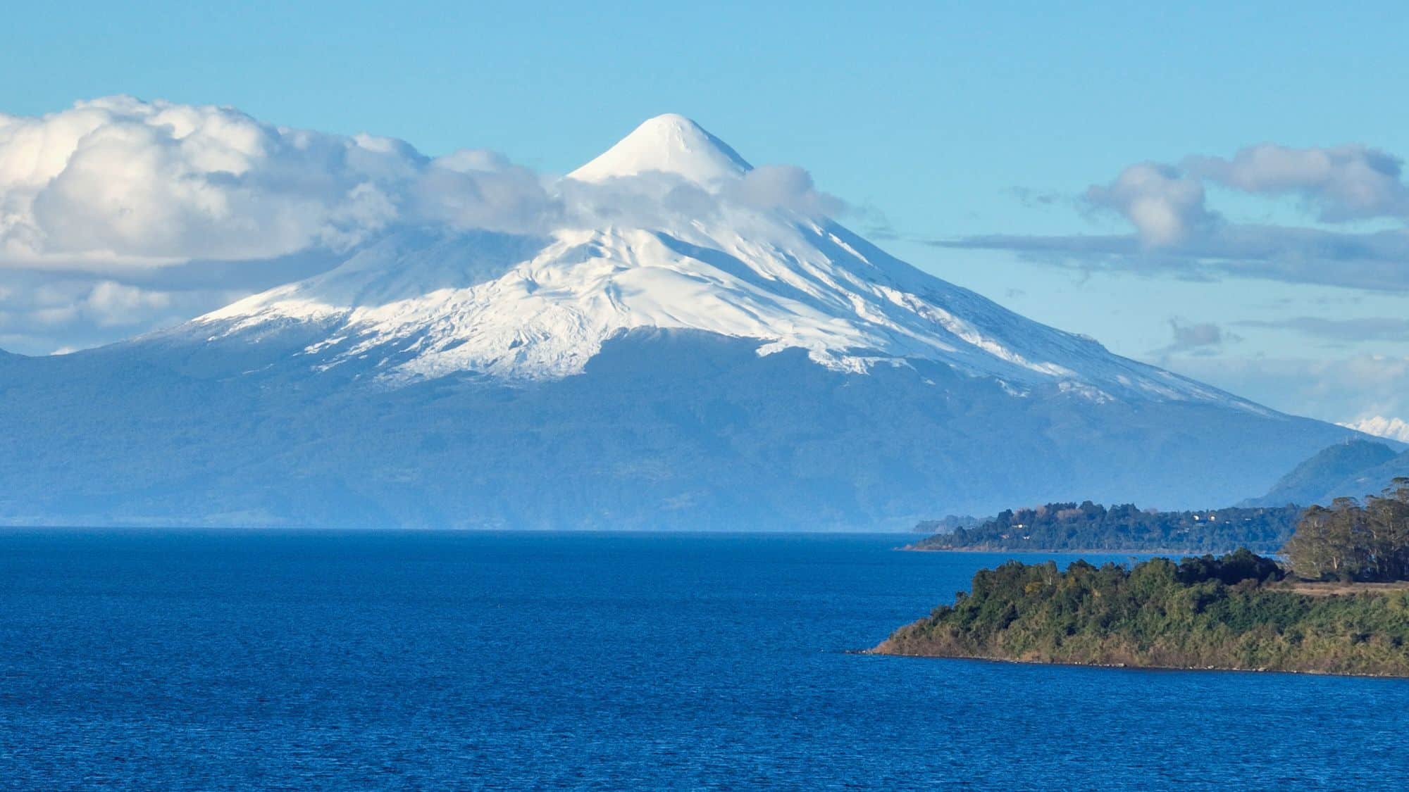 A snowy volcanic peak rises dramatically in the distance beyond a large deep blue lake, with a forested shoreline in the foreground.