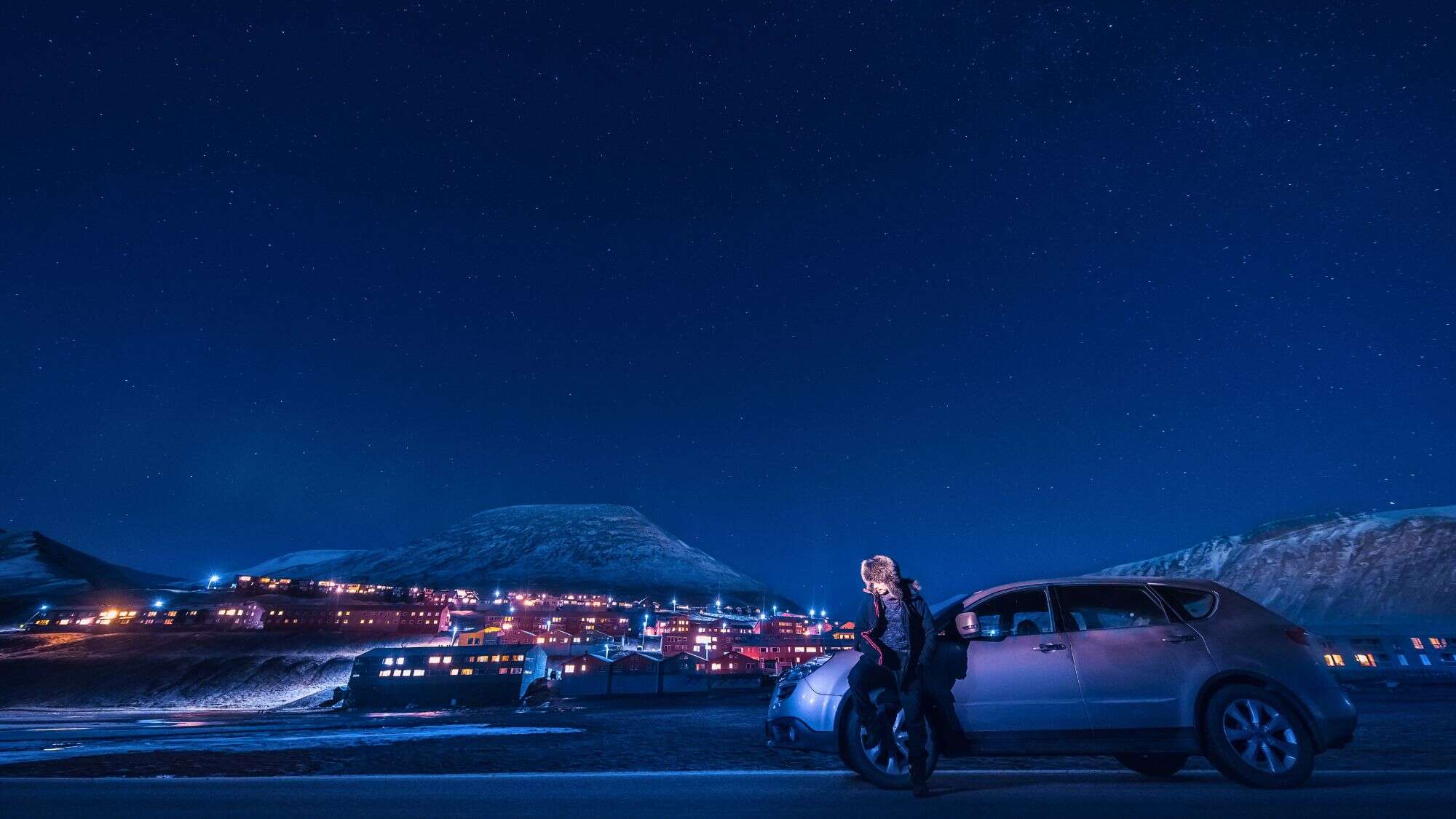 A person stands by a frosty car under a deep starry sky, with the warmly lit town of Longyearbyen and snow-covered hills glowing in the Arctic night.