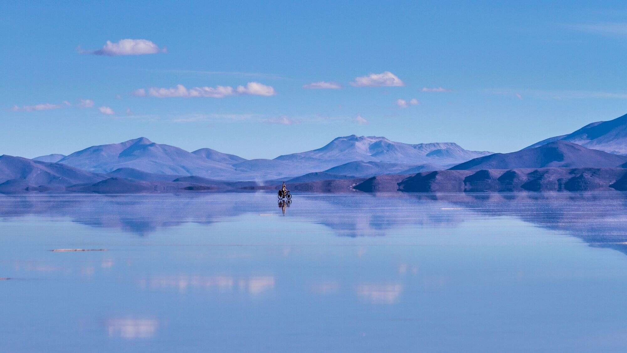 A lone cyclist rides across a gleaming mirror of water on Bolivia’s Uyuni Salt Flats, with distant blue mountains reflected in the horizon.