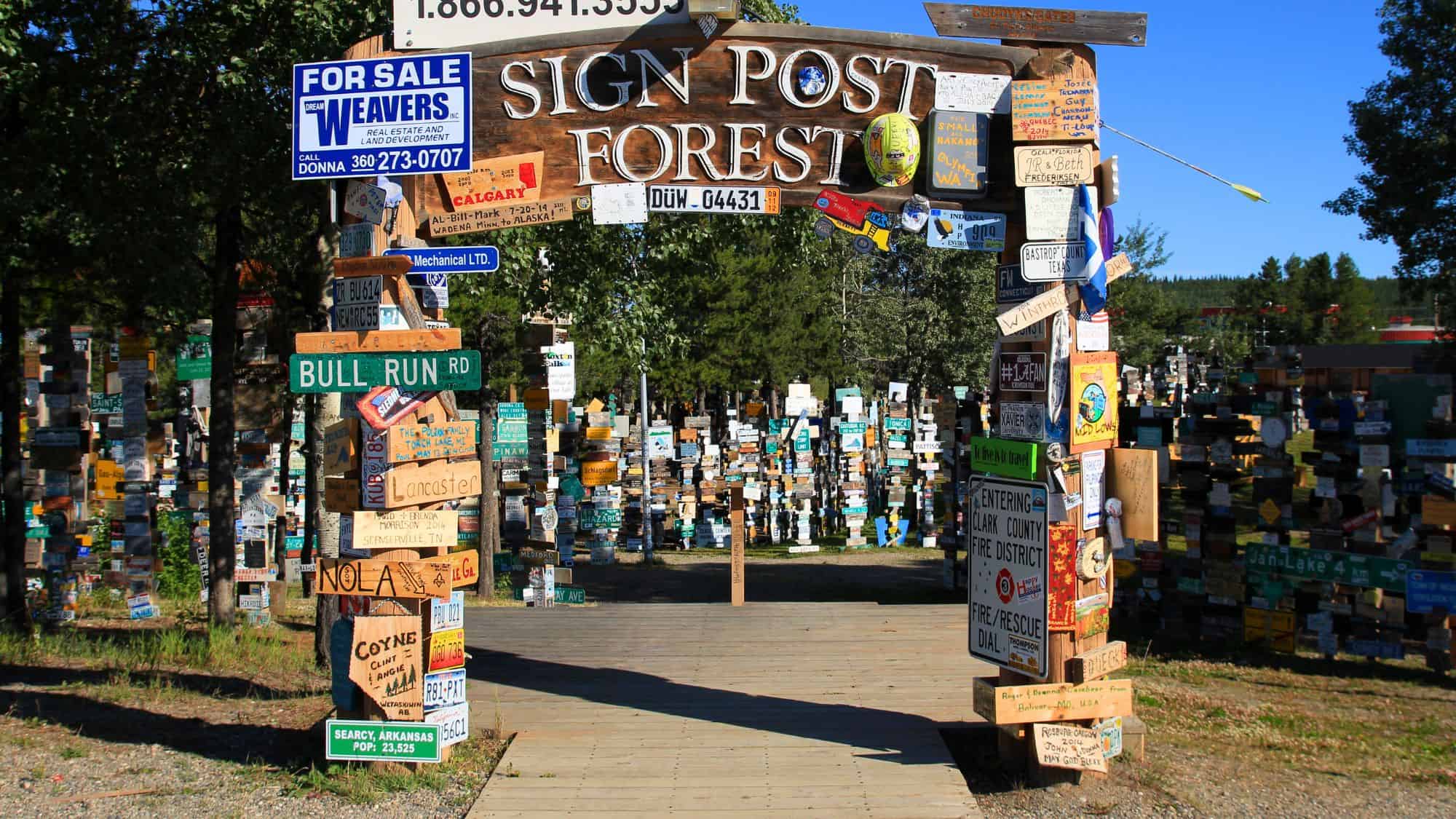 An eclectic forest of signs in Watson Lake, Yukon, with thousands of license plates, street signs, and handmade markers from around the world, all clustered under the entrance arch labeled “Sign Post Forest.”